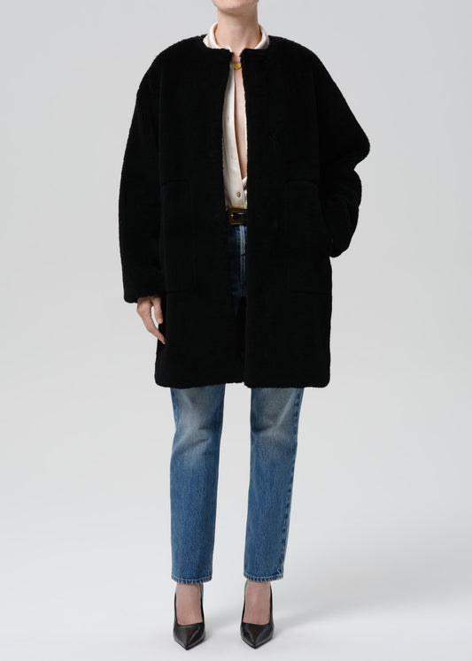 A model wearing a black oversized teddy coat over a cream blouse, blue jeans, and black pointed heels, photographed against a grey background.