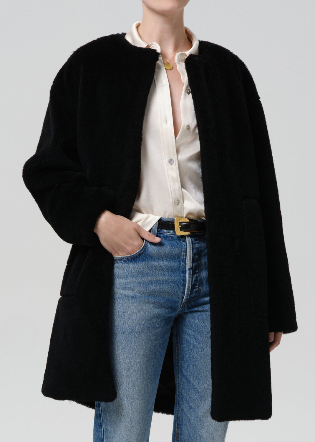 A model wearing a black fuzzy mid-length coat over a cream button-up shirt and blue jeans with a gold-buckled belt, photographed against a grey background.