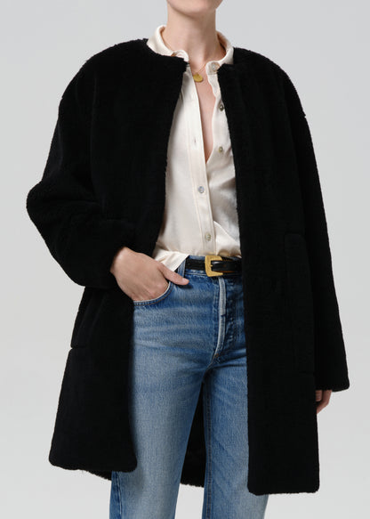 A model wearing a black fuzzy mid-length coat over a cream button-up shirt and blue jeans with a gold-buckled belt, photographed against a grey background.