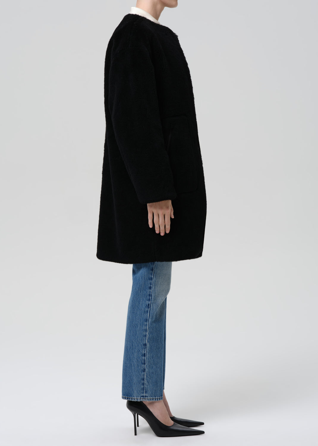 A model shown from the side wearing a black textured coat, blue jeans, and black high heels against a grey background.