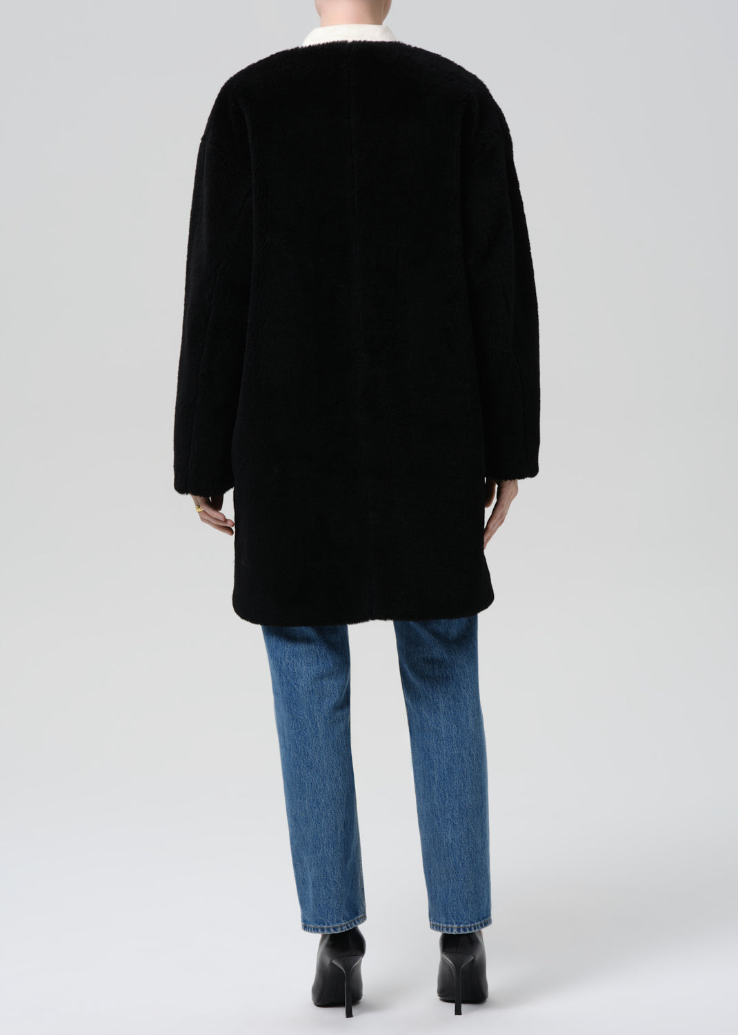 A model shown from the back wearing a black mid-length coat, blue jeans, and black high-heeled boots against a grey background.
