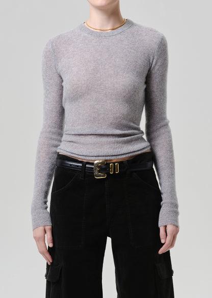 A model wearing a light grey ribbed knit crewneck sweater, paired with black corduroy trousers and a black belt with a gold buckle, against a grey background.