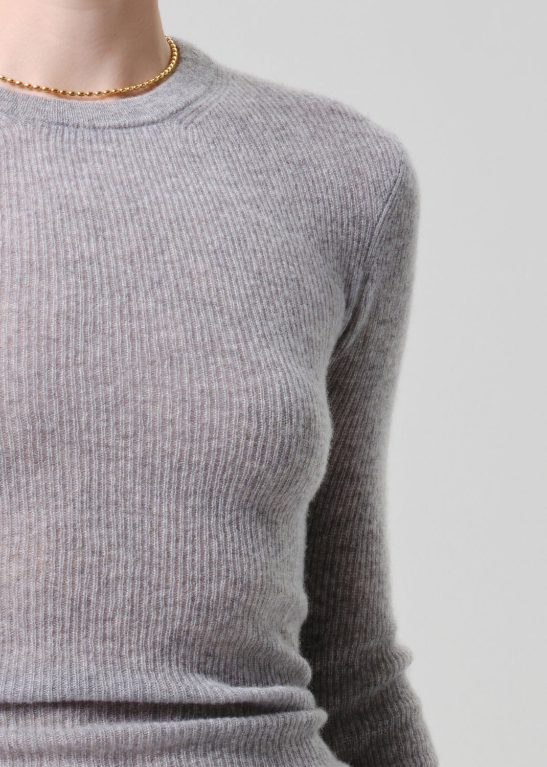 Close-up of a person wearing a light grey ribbed knit crewneck sweater, paired with a delicate gold beaded necklace, against a light grey background.