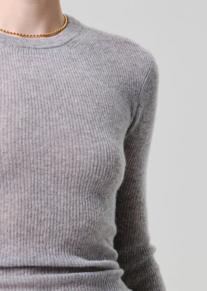 Close-up of a person wearing a light grey ribbed knit crewneck sweater, paired with a delicate gold beaded necklace, against a light grey background.