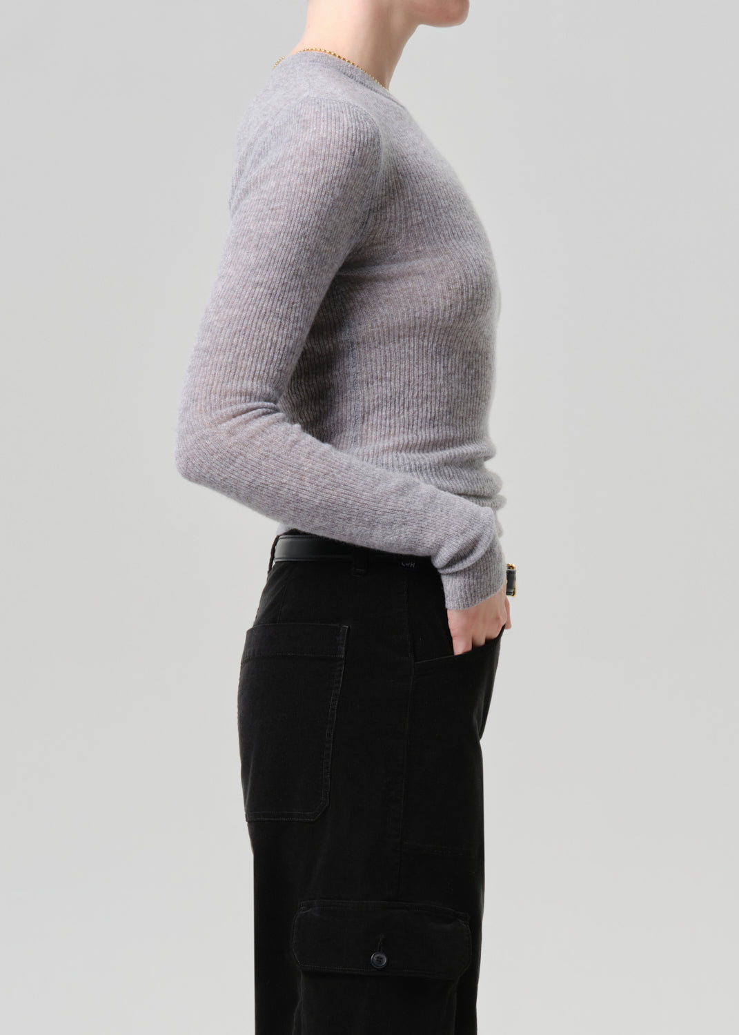 A side-profile view of a person wearing a light grey ribbed knit long-sleeve sweater and black cargo pants against a grey background.