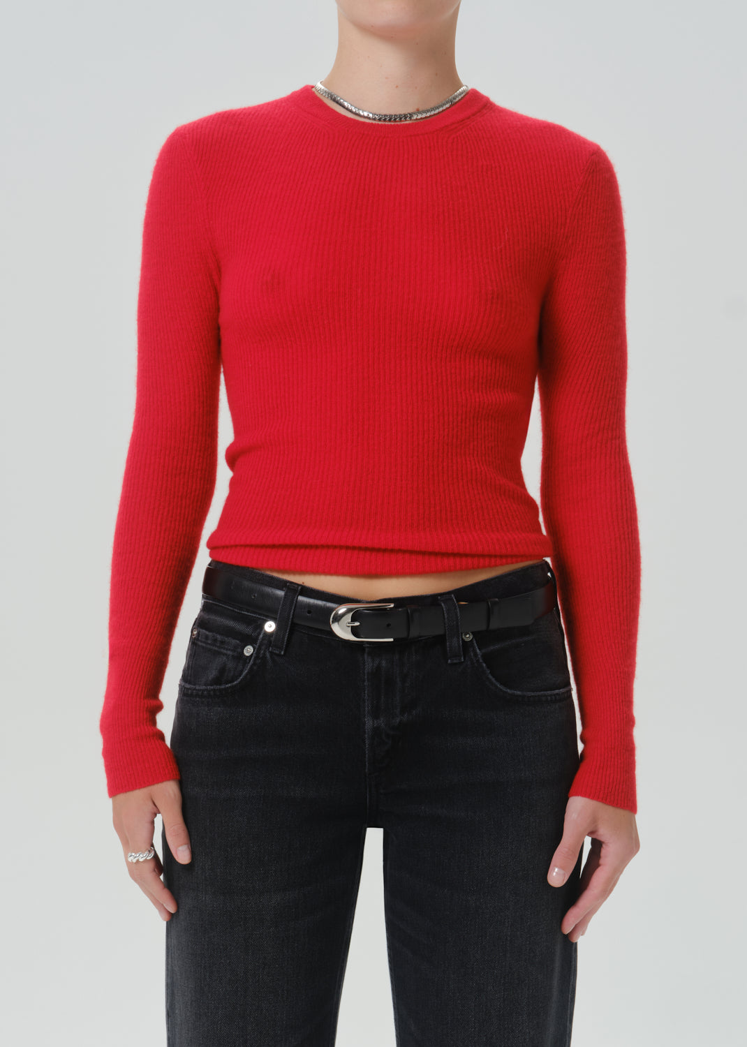 A model wearing a bright red ribbed knit long-sleeve crop top with a silver chain detail at the neckline, paired with black jeans and a black belt, against a light grey background.