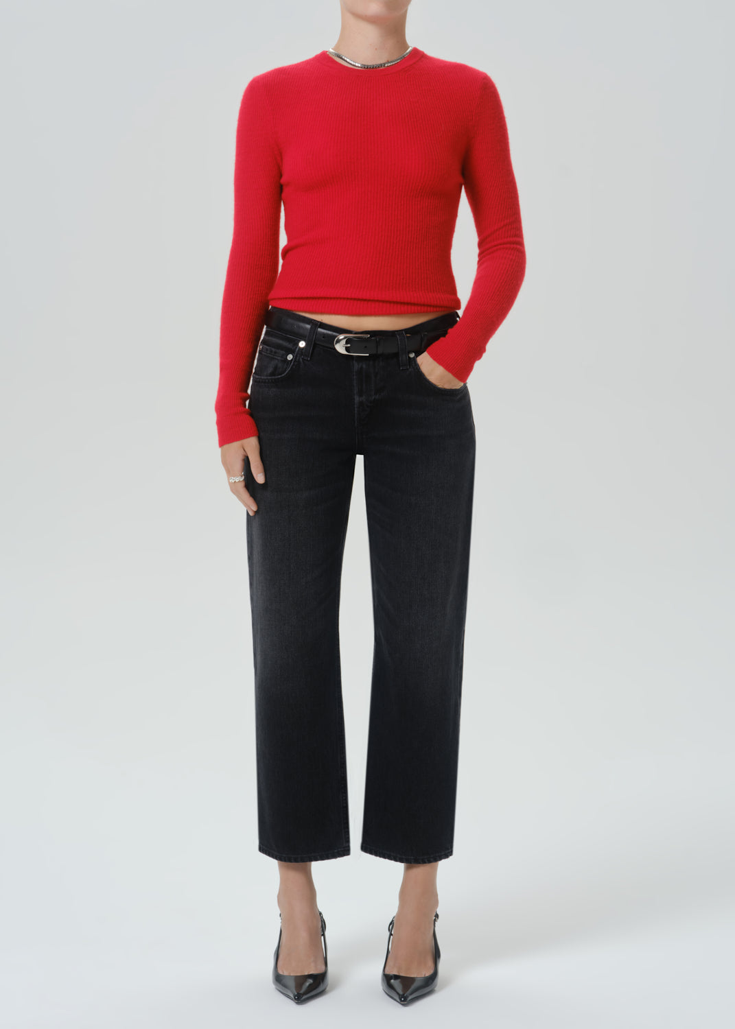 A model wearing a bright red long-sleeve crewneck top, black cropped wide-leg jeans with a black belt, and black pointed-toe heels, photographed against a grey background.