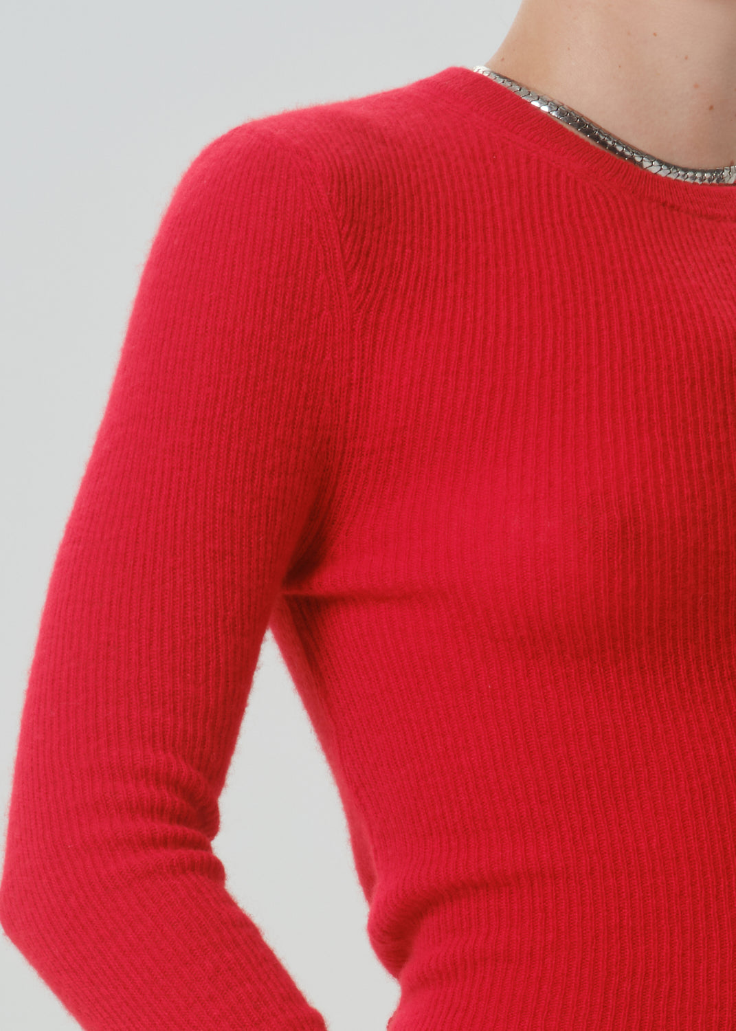 A close-up of a person wearing a bright red ribbed knit sweater with a silver rhinestone-trimmed crew neckline, set against a grey background.