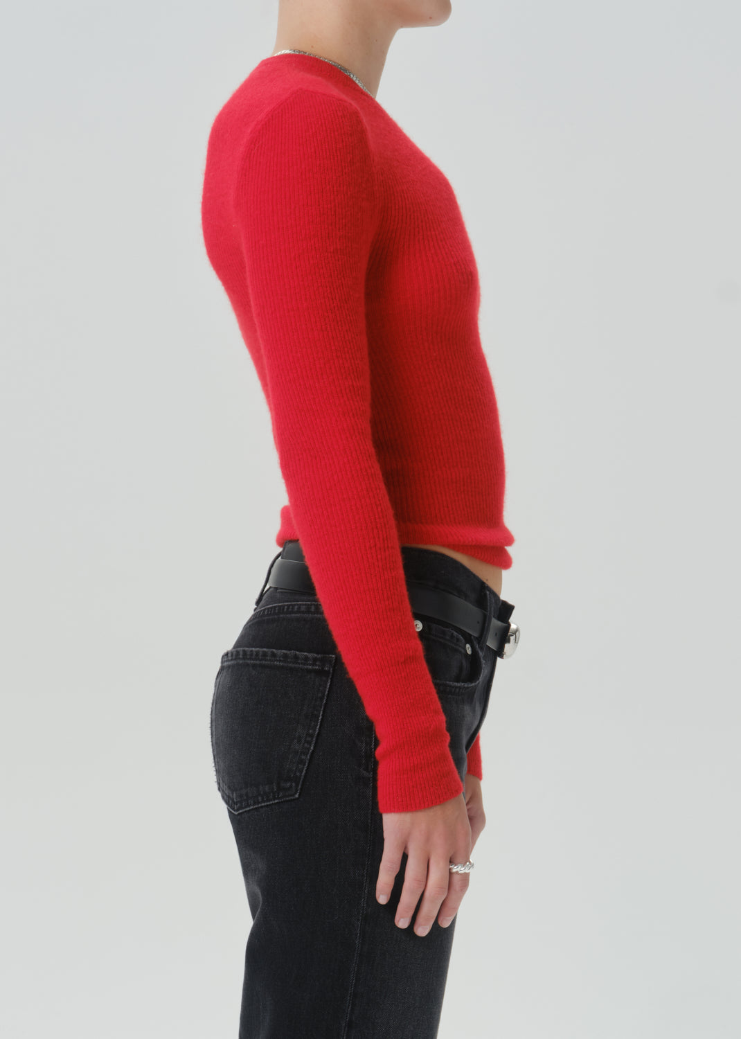 A side-profile view of a model wearing a vibrant red long-sleeve knit sweater and black jeans with a black belt, set against a grey background.