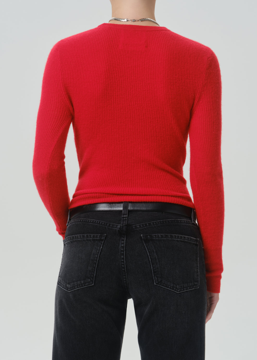 A person shown from the back wearing a bright red ribbed long-sleeve sweater and dark jeans against a grey background.