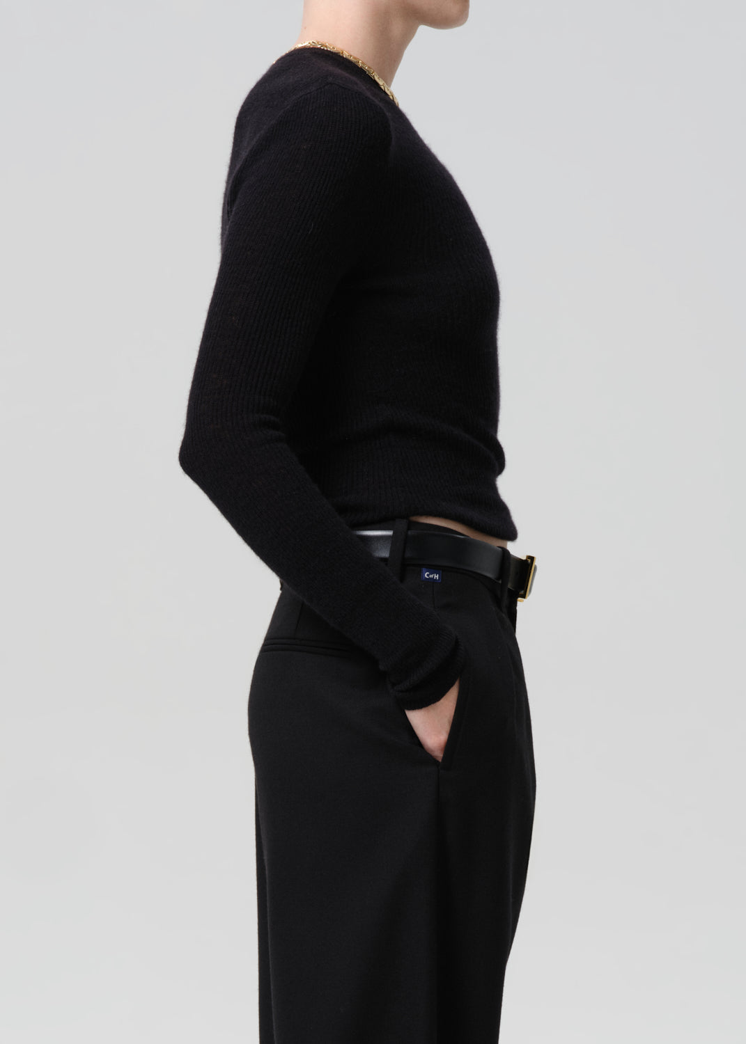 Side of person wearing black sweater with black belt and black pants on a grey background