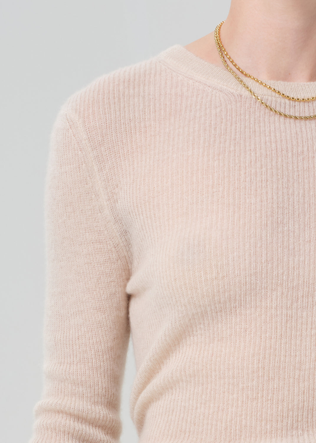 Close-up of a person wearing a light cream ribbed sweater and gold necklaces on a gray background
