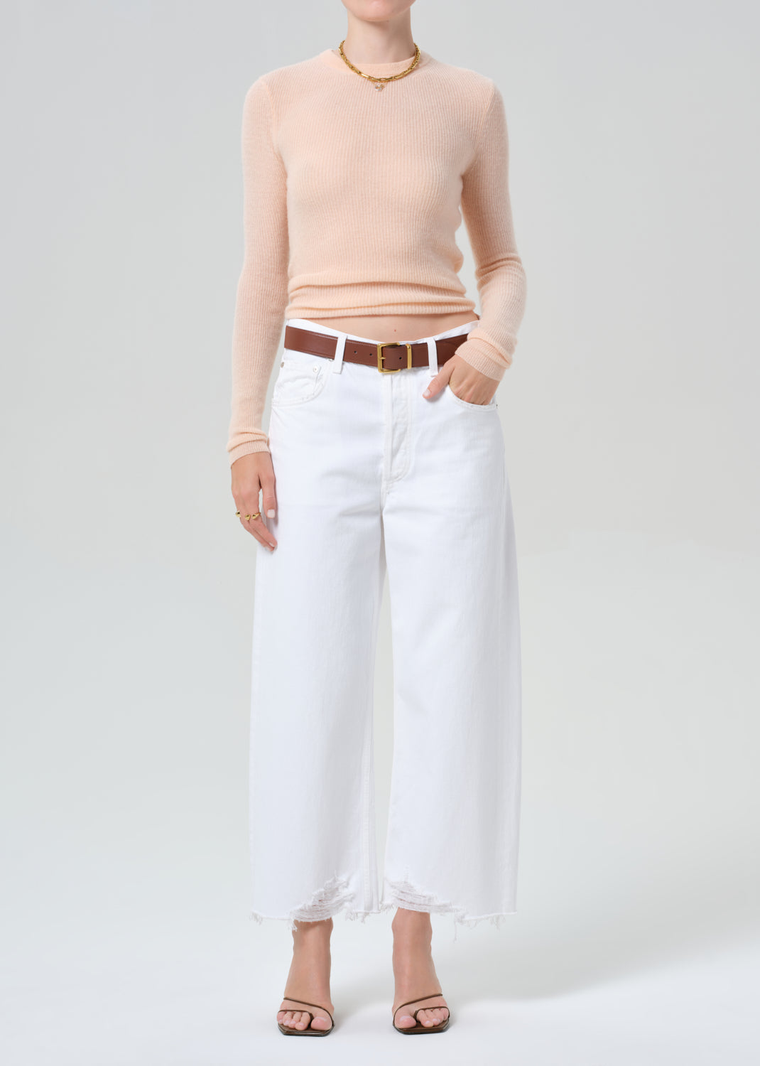Person wearing a light peach sweater and white jeans with a brown belt on a plain background
