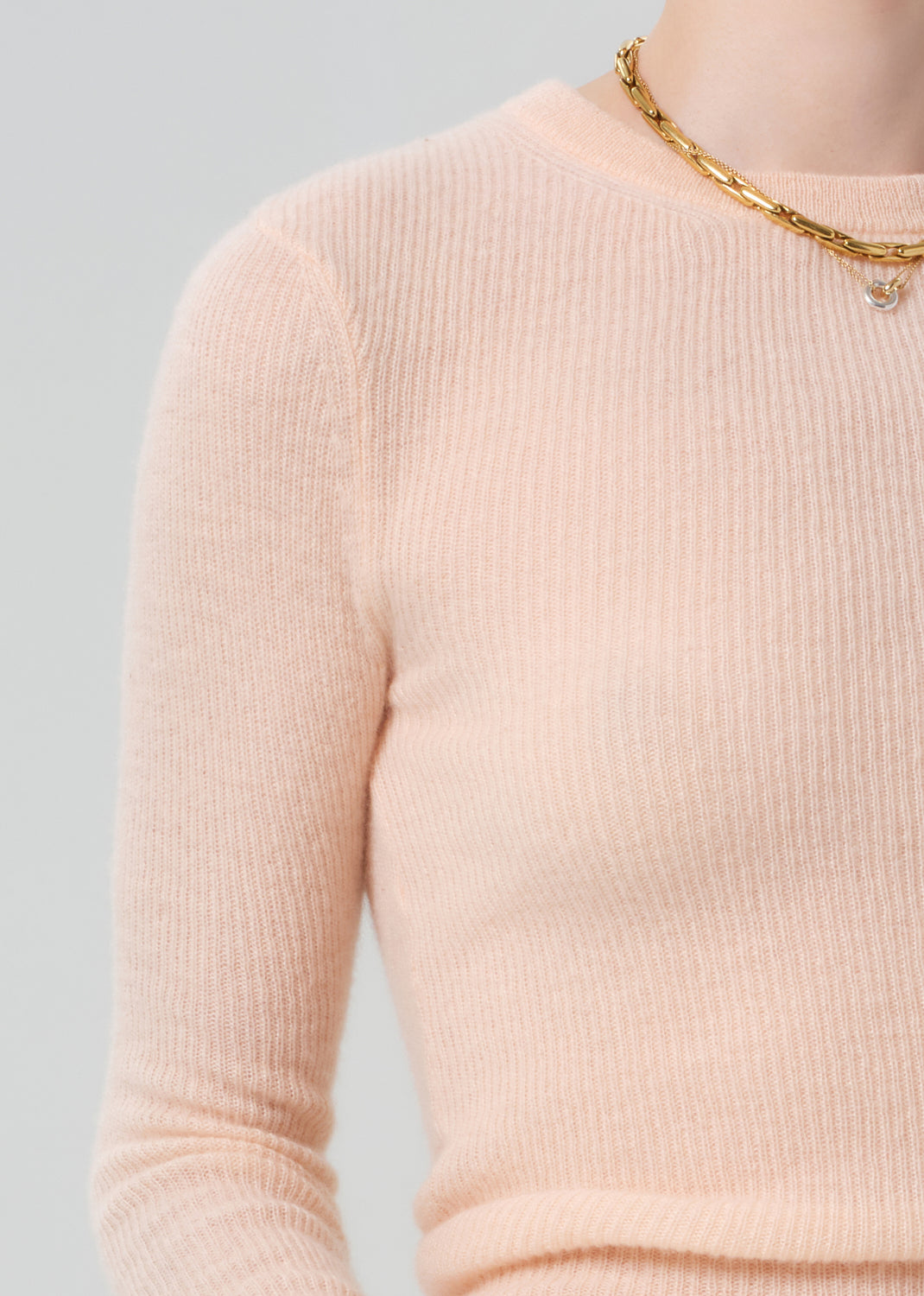 Person wearing a light peach ribbed sweater with a gold necklace on a gray background