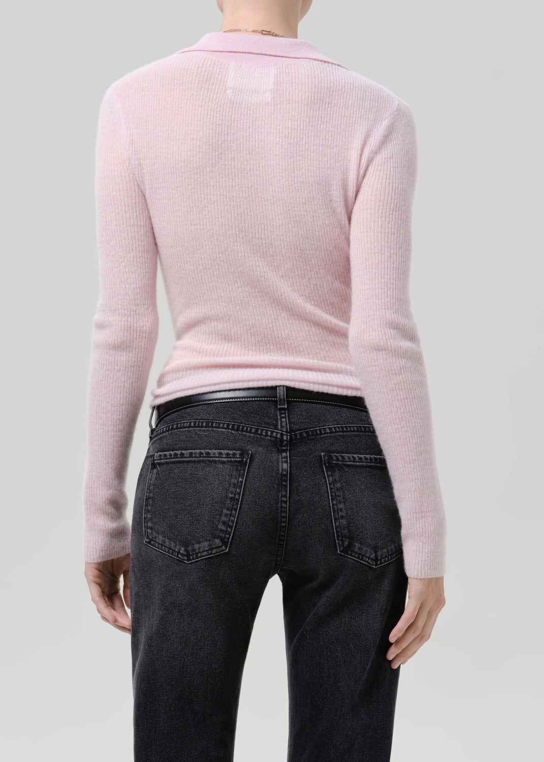 Person wearing a light pink ribbed knit polo sweater with black jeans viewed from the back on a grey background