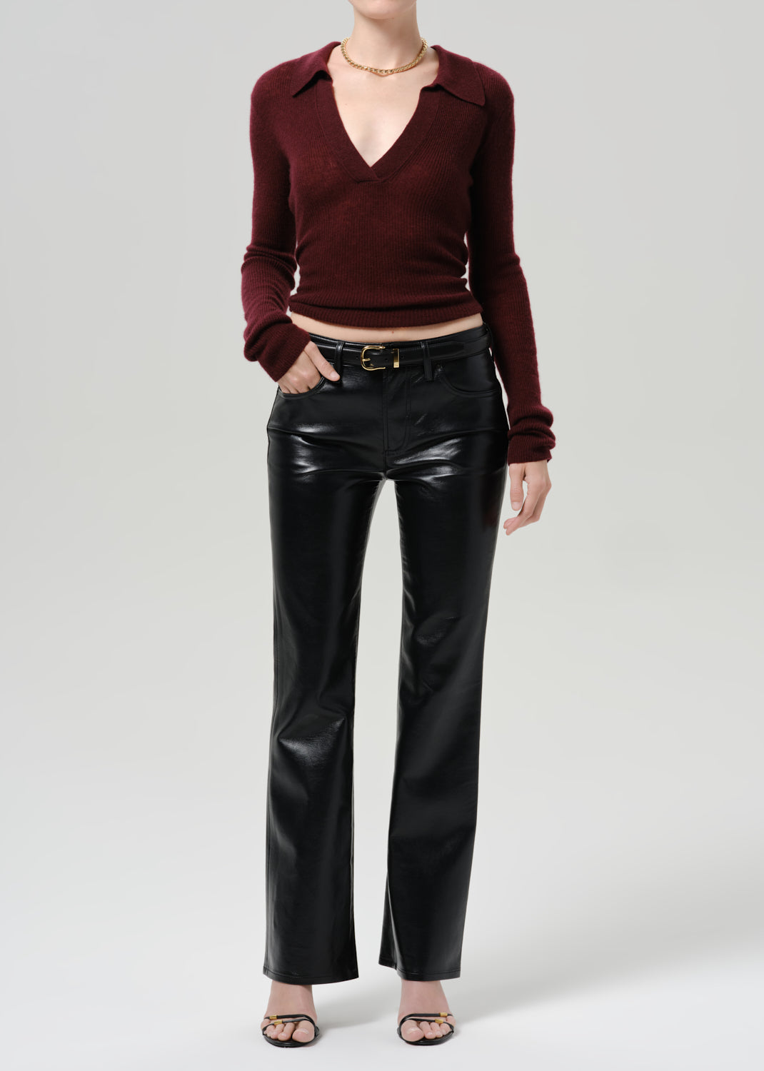 A model wearing a dark burgundy long-sleeve knit polo-collar top and shiny black faux-leather flare pants, styled with a gold belt and black sandals against a grey background.