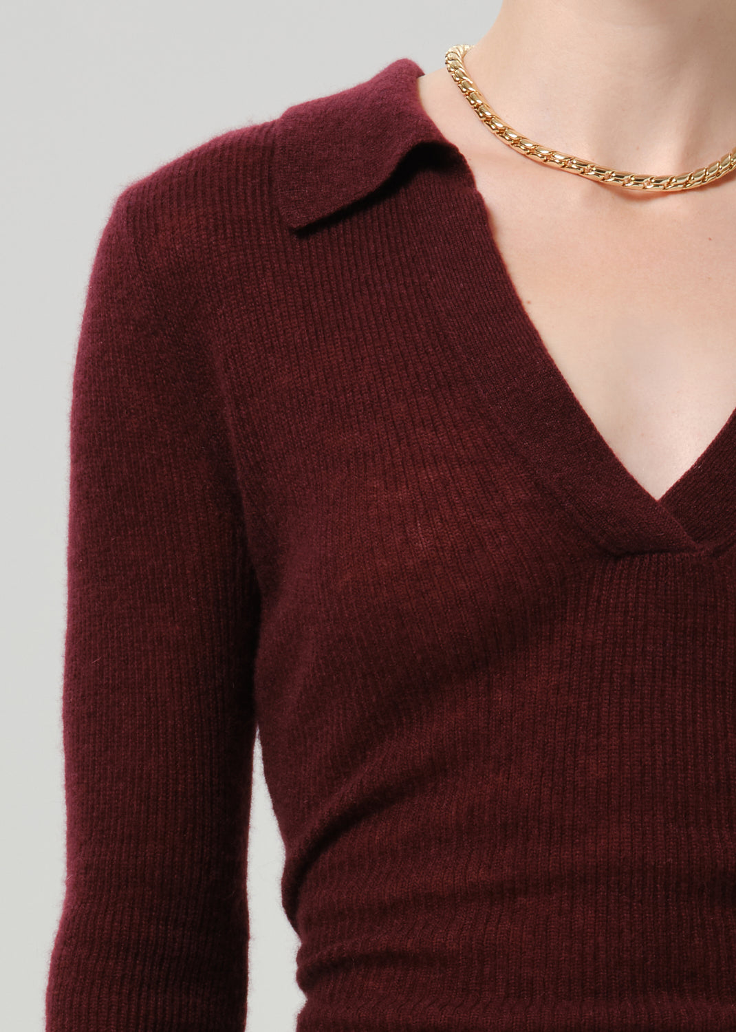 A close-up of a person wearing a ribbed knit long-sleeve polo-style top in deep burgundy, paired with a gold chain necklace, against a grey background.