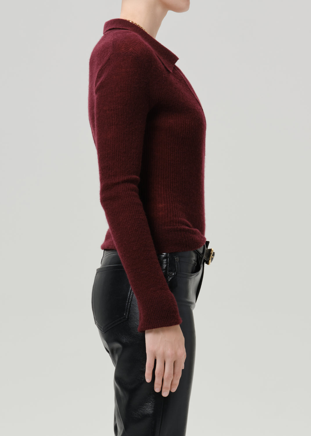 A side profile view of a model wearing a ribbed knit polo sweater in deep burgundy and black leather pants, set against a grey background.
