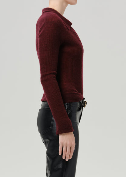 A side profile view of a model wearing a ribbed knit polo sweater in deep burgundy and black leather pants, set against a grey background.