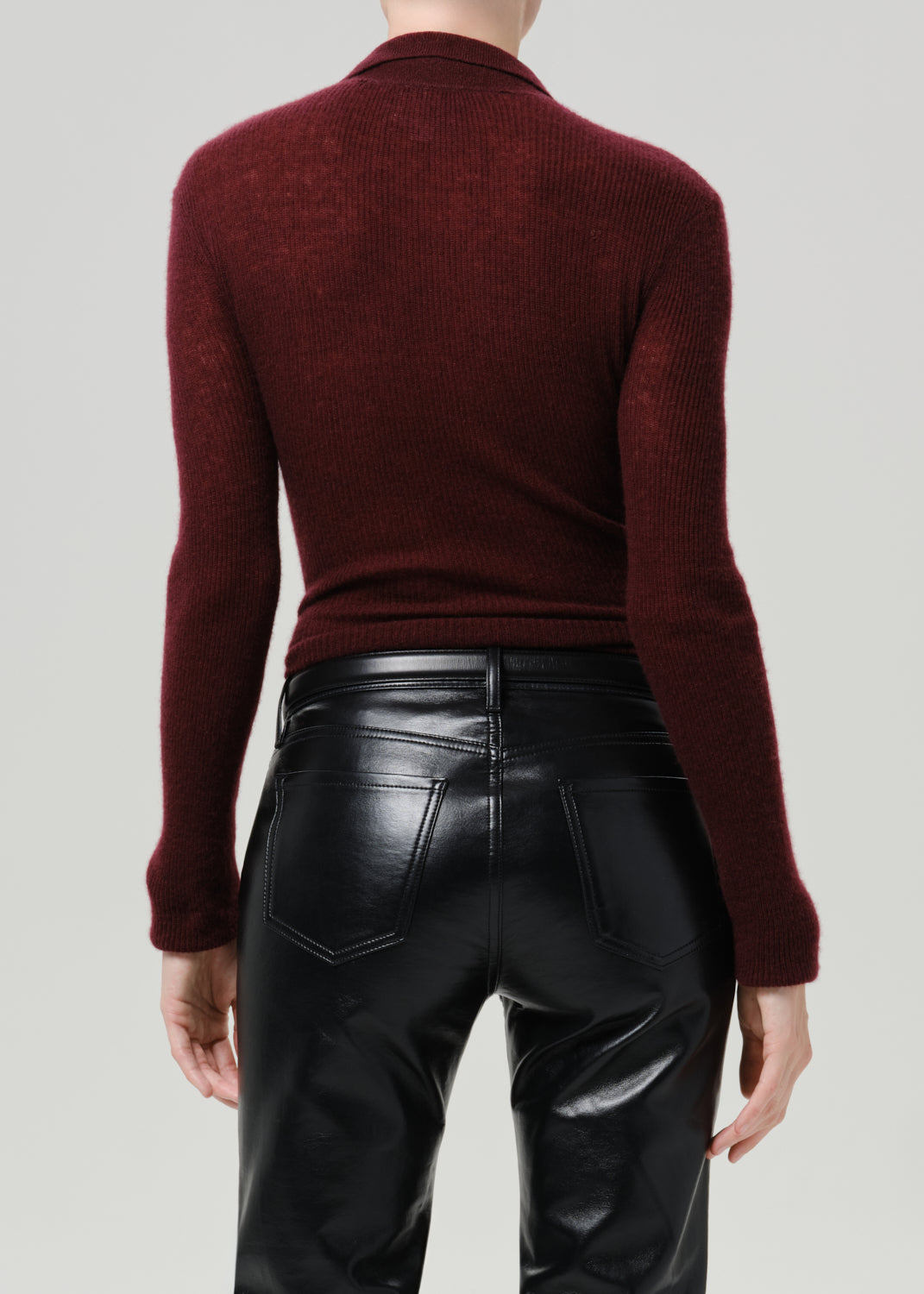Rear view of a model wearing a ribbed long-sleeve mock neck sweater in deep burgundy and black faux-leather pants, against a grey background.