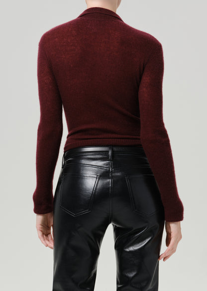 Rear view of a model wearing a ribbed long-sleeve mock neck sweater in deep burgundy and black faux-leather pants, against a grey background.