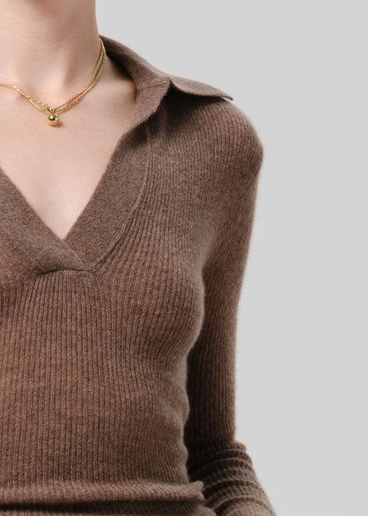 A close-up of a person wearing a ribbed brown knit sweater with a V-neck and collar, paired with a delicate gold necklace, against a grey background.