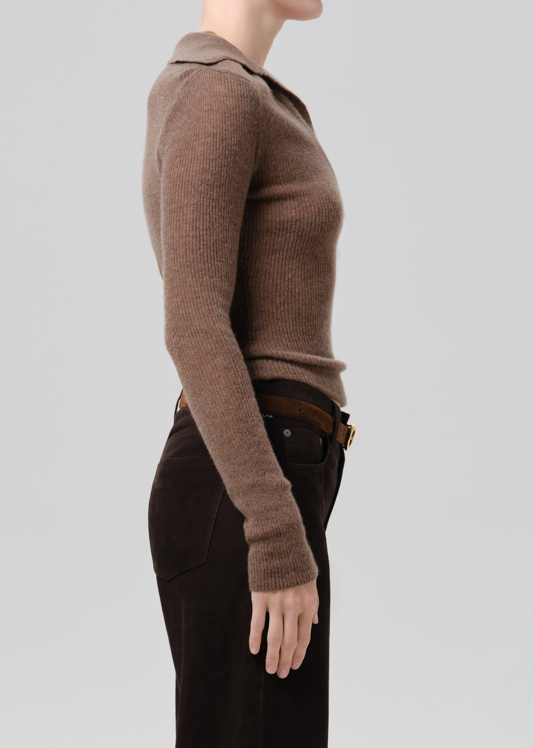 A side-profile view of a model wearing a ribbed knit long-sleeve polo-style sweater in taupe brown, paired with dark brown trousers and a brown belt, against a grey background.