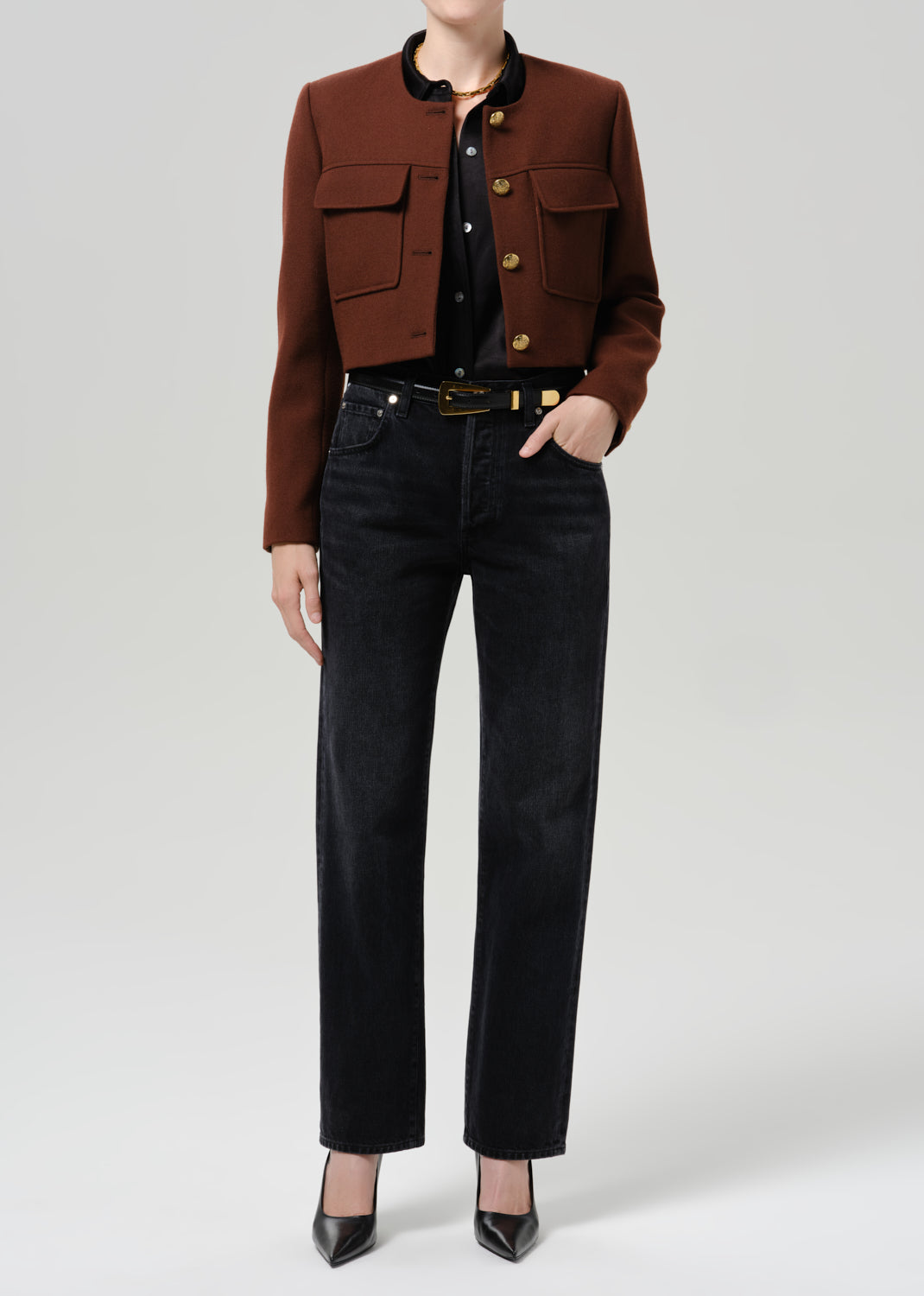 Person wearing a brown tailored cropped jacket with black straight leg jeans on a grey background