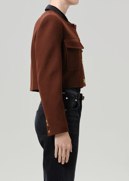 Person wearing a brown tailored cropped jacket with black straight leg jeans viewed from the side on a grey background