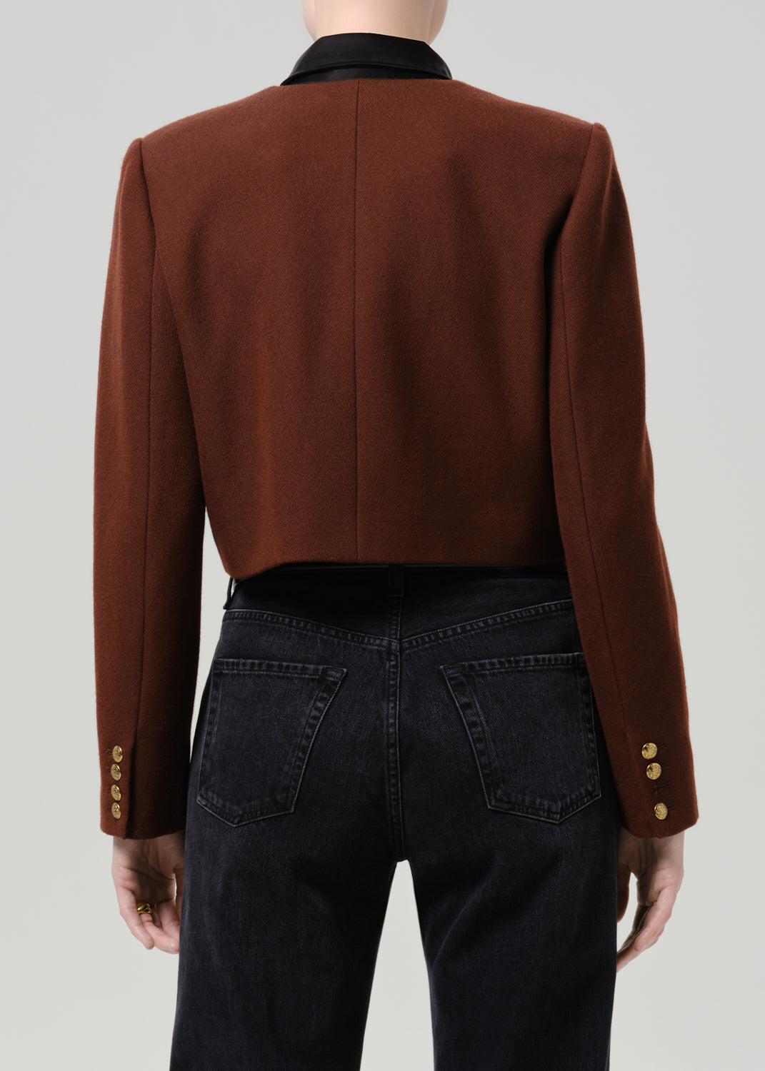 Person wearing a brown tailored cropped jacket with black straight leg jeans viewed from the back on a grey background