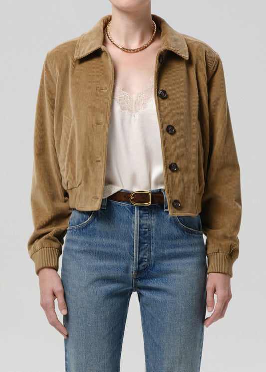 A cropped brown corduroy jacket worn by a model, styled with a cream lace-trim camisole, blue jeans, and a brown belt against a grey background.