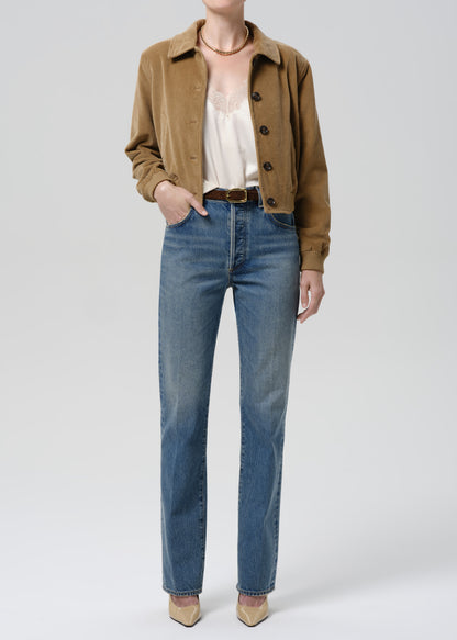 Person wearing a brown jacket, white shirt, and blue jeans on a grey background.