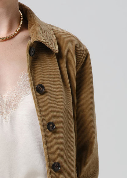 Close-up of a person wearing a brown corduroy button-front jacket over a white lace-trim top, with a gold chain necklace visible against a grey background.