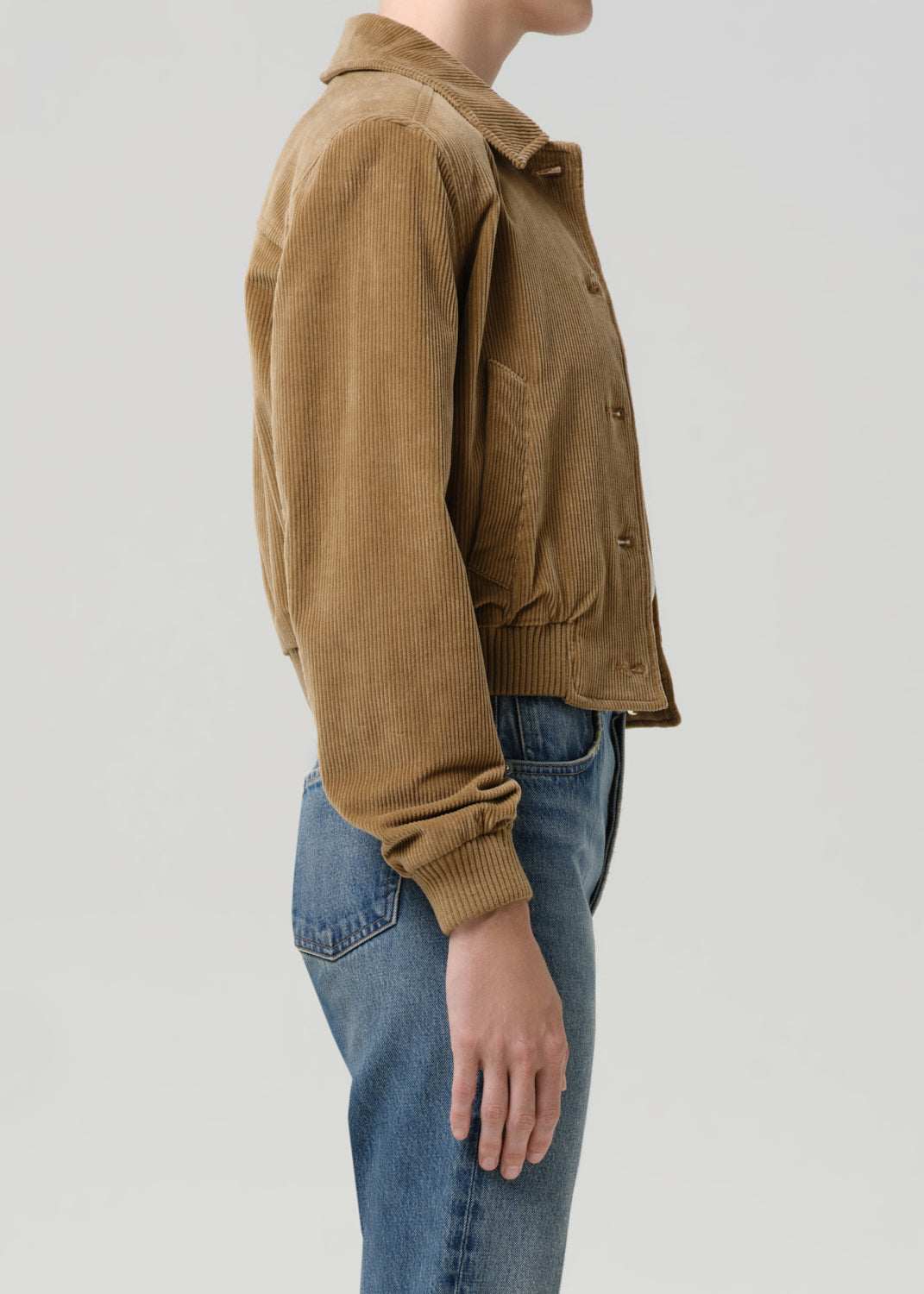A side-profile view of a person wearing a cropped tan corduroy jacket with ribbed collar, cuffs, and hem, paired with blue jeans, against a grey background.