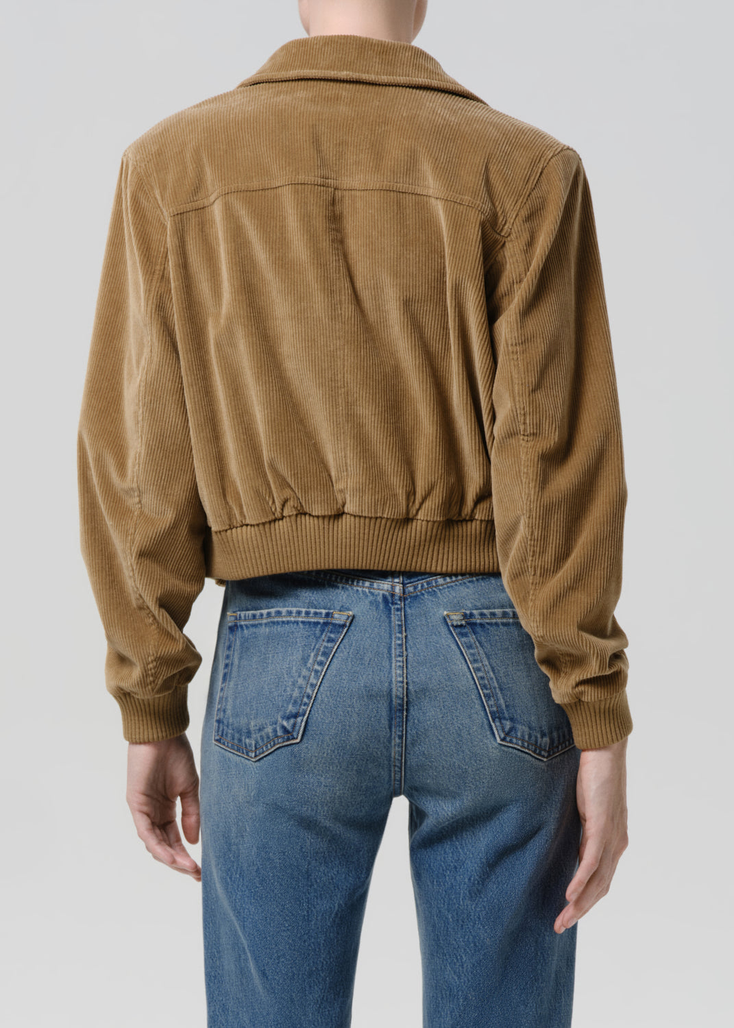 A person shown from the back wearing a cropped tan corduroy jacket with a ribbed hem and cuffs, paired with blue jeans, against a grey background.