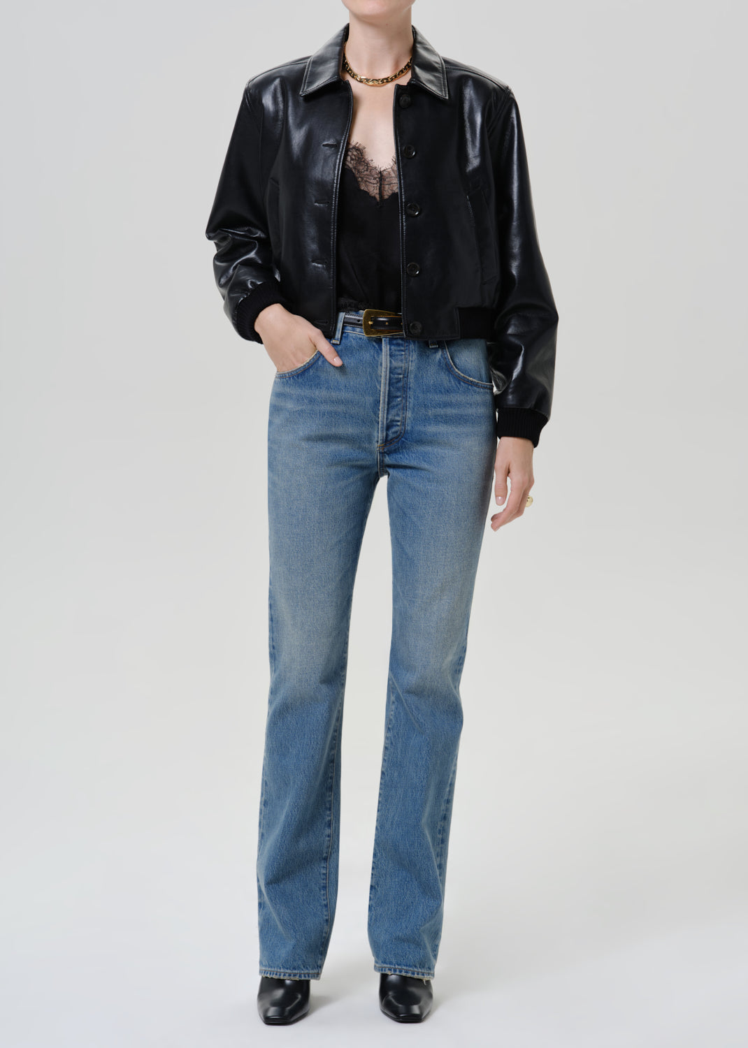 Woman wearing a black leather button-up shirt with medium blue straight leg jeans on a grey background.