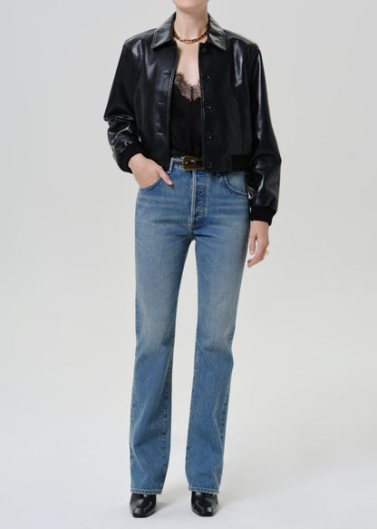 Woman wearing a black leather button-up shirt with medium blue straight leg jeans on a grey background.