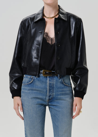 A model wearing a black faux-leather cropped jacket with a collar and button front, paired with blue jeans and a black lace top, against a light gray studio background.