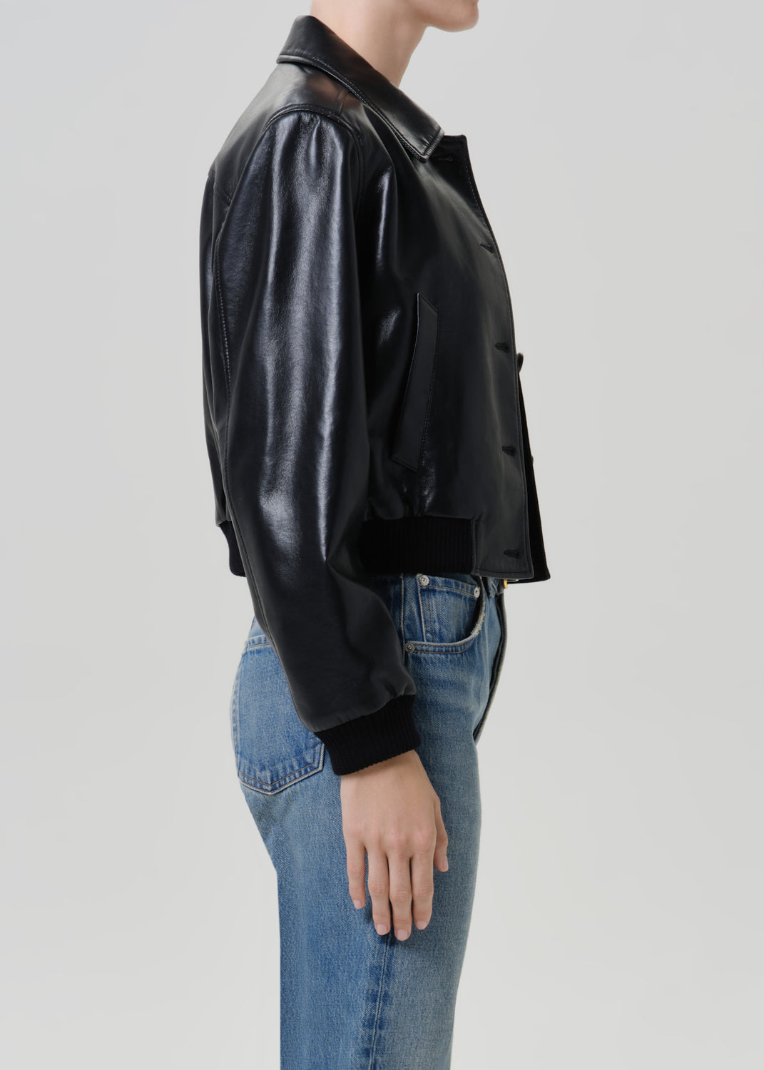 Woman wearing a black leather button-up shirt with medium blue straight leg jeans viewed from the side on a grey background.