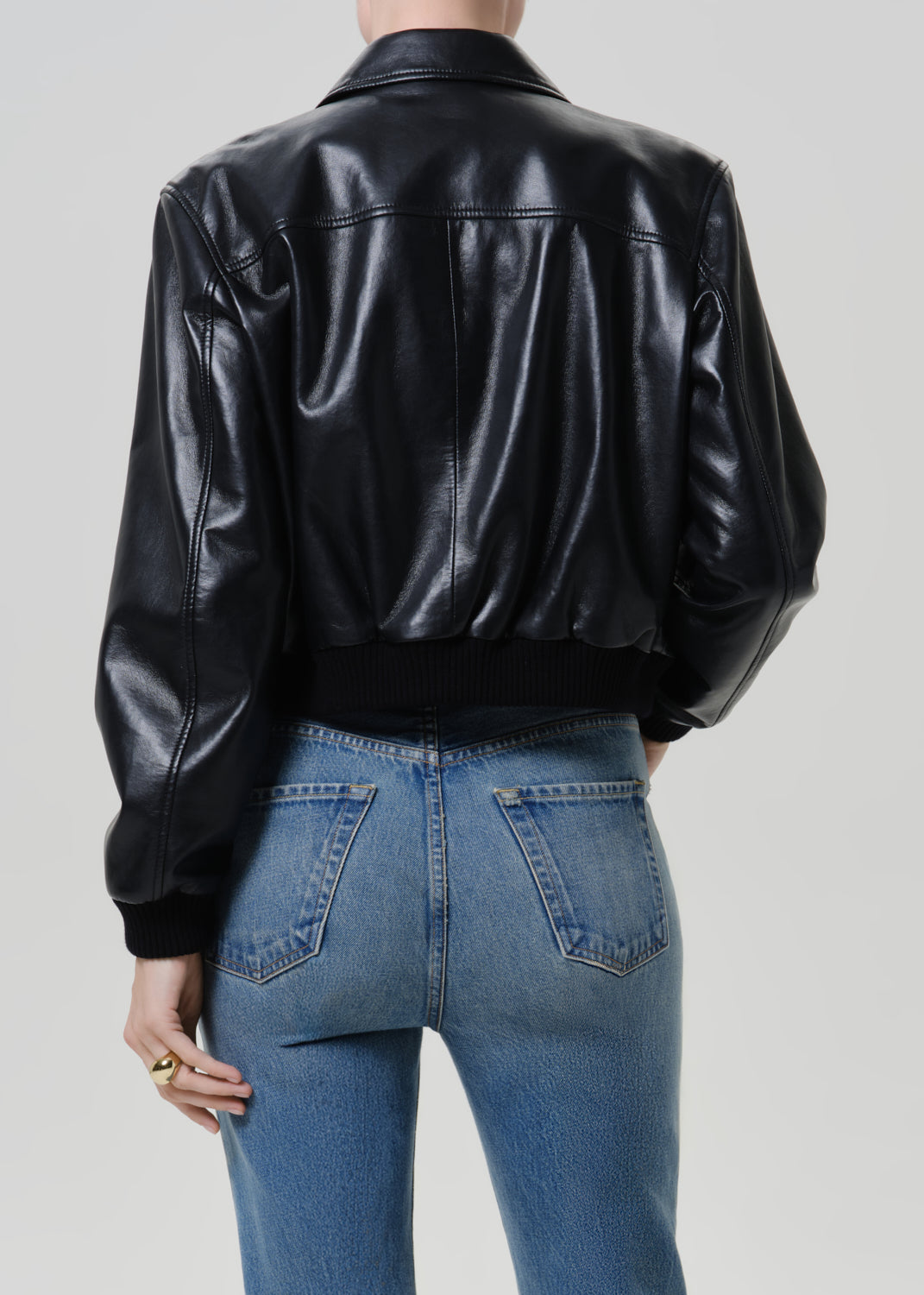 Woman wearing a black leather button-up shirt with medium blue straight leg jeans viewed from the back on a grey background.