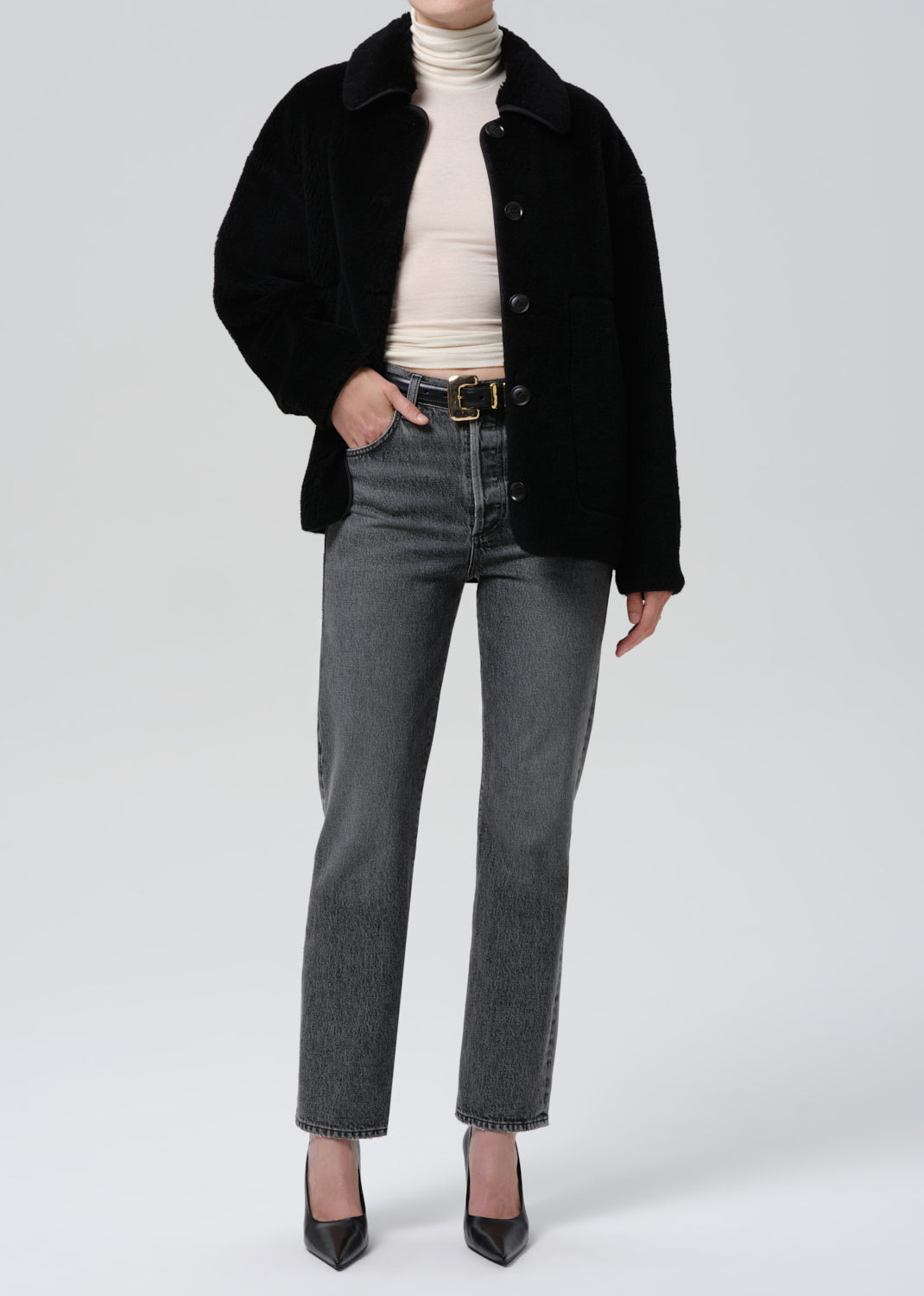Person wearing a black coat, beige turtleneck, gray jeans, and black shoes on a grey background