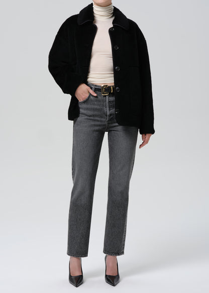 Person wearing a black coat, beige turtleneck, gray jeans, and black shoes on a grey background