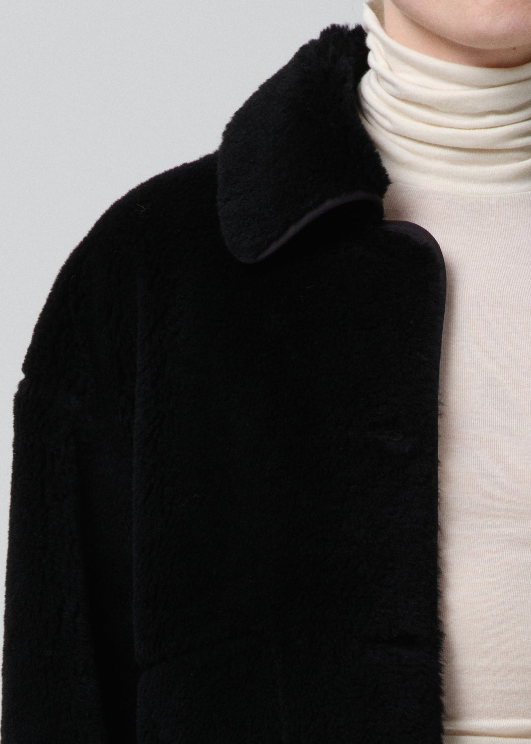 Black coat with a textured collar worn over a white turtleneck sweater on a grey background