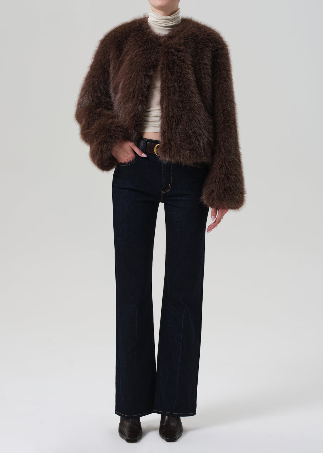 Person wearing a brown fur coat, white turtleneck, black pants, and shoes on a light gray background