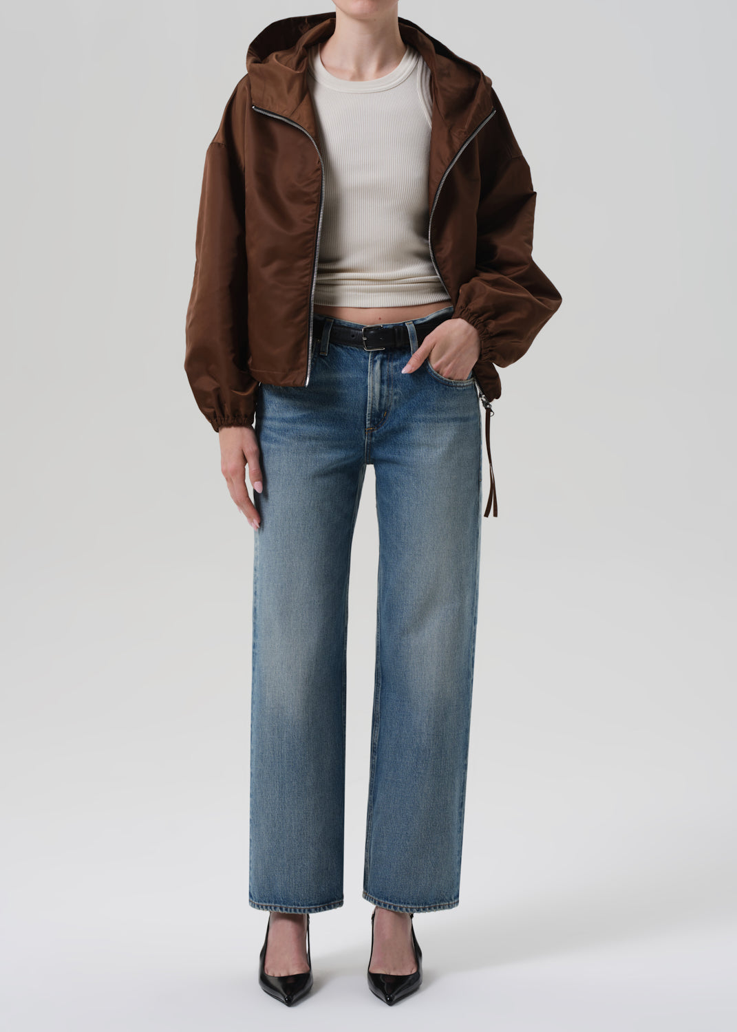 Person wearing a brown jacket, white top, and blue jeans on a white background