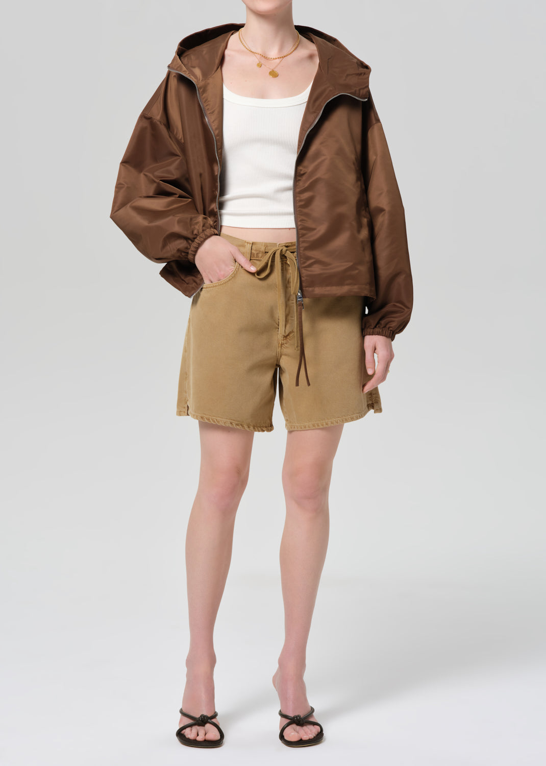 Person wearing a brown jacket, white top, and beige shorts on a plain background