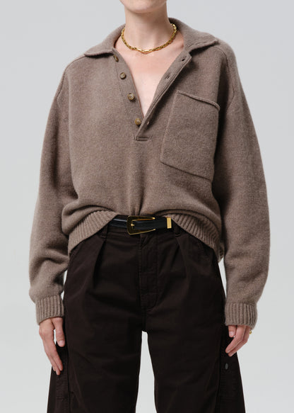 Person wearing a brown sweater with a high collar and black pants on a grey background