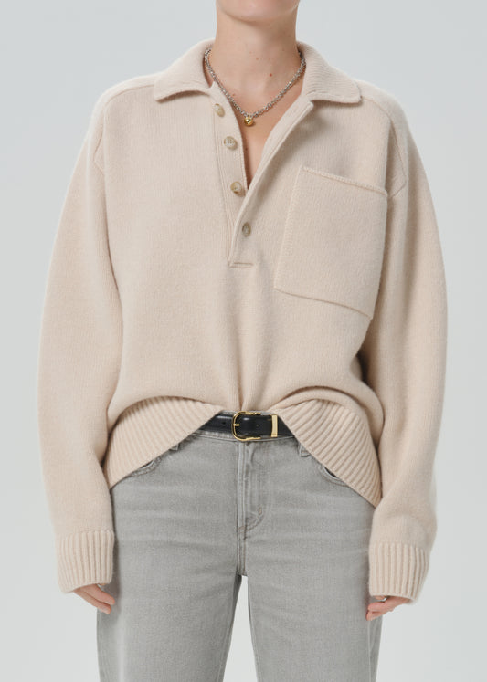 A person wearing a light beige knit polo sweater with a collar, three-button placket, and chest pocket, paired with light grey jeans and a black belt, against a grey background.