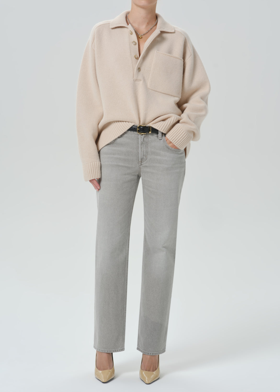 A model wearing a light beige knit polo sweater, light grey straight-leg jeans, a black belt, and beige pointed-toe heels, photographed against a seamless grey background.
