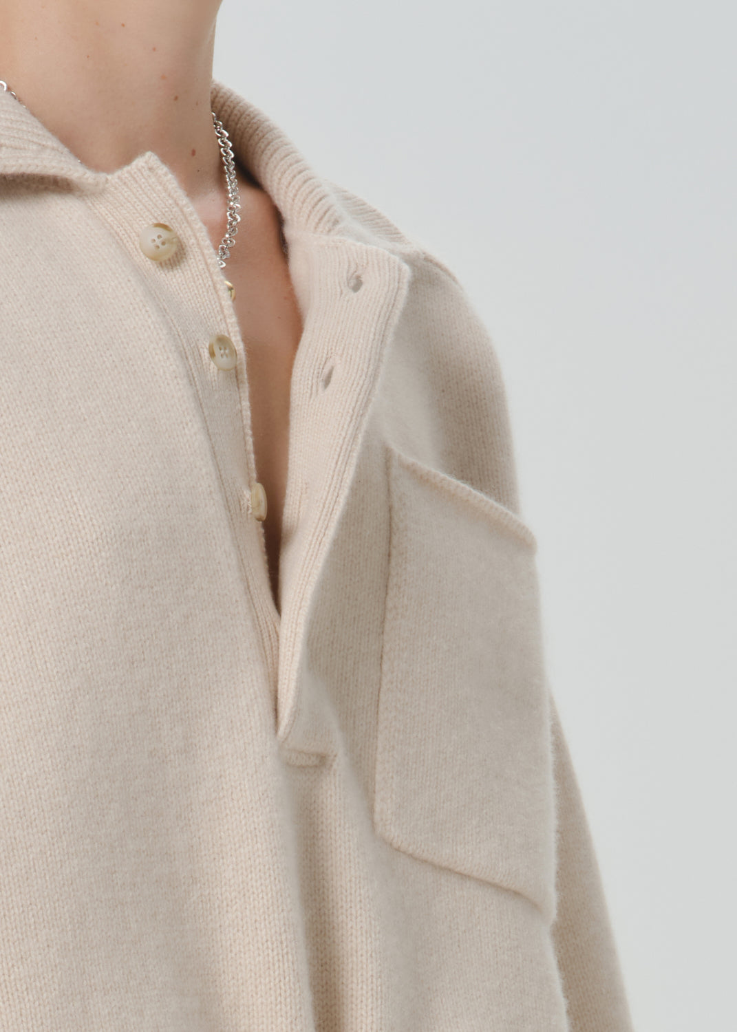 Close-up of a person wearing a soft knit polo-style sweater in a light warm beige tone, with a collar, partial button placket, and a large chest pocket against a grey background.
